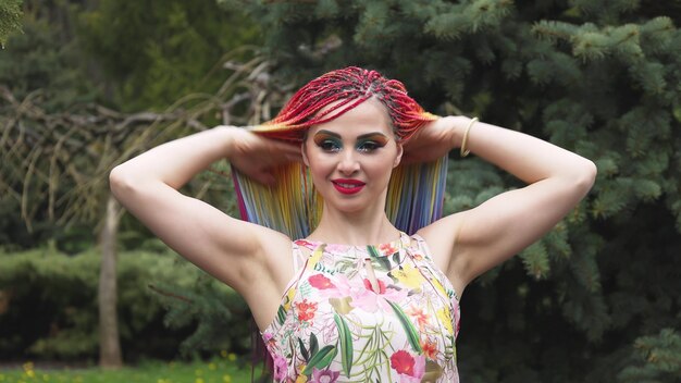 Garota feliz em um vestido com tranças africanas de arco-íris e maquiagem colorida brilhante. uma fofa sorri e joga o cabelo para cima e gira em uma caminhada no parque da primavera