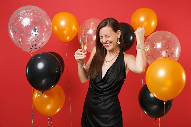 Garota feliz em um vestido preto comemorando, segurando a taça de champanhe, cerrando os punhos como vencedor em balões de ar de fundo vermelho brilhante. dia da mulher, feliz ano novo, conceito de festa de feriado de maquete de aniversário.