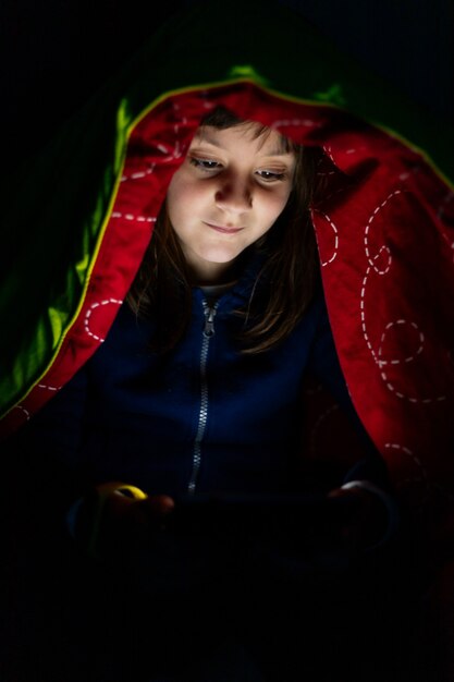 Garota feliz enquanto assistia ao Tablet, em seu quarto com um cobertor por cima