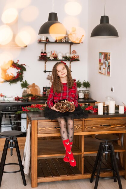 Foto garota feliz está sentado na cozinha com uma guirlanda de natal. com roupas xadrez vermelhas e um laço vermelho no cabelo. casa de ano novo