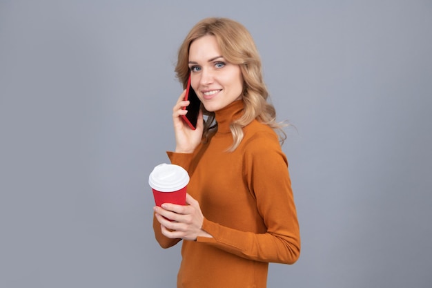 Garota feliz fala no celular segurando uma xícara quente de chá ou café bebendo fundo cinza, pausa.