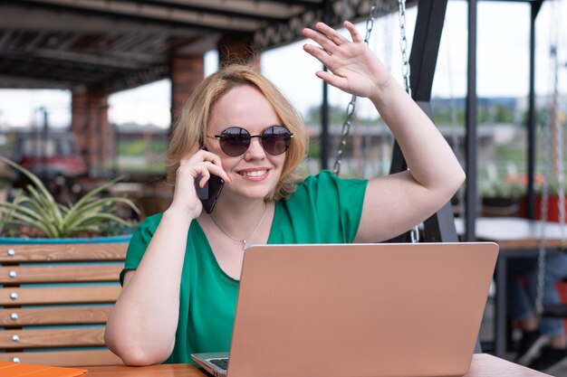 Garota feliz falando ao telefone com amigos navegando no laptop e acenando com a mão cumprimentando amigos em um café de rua