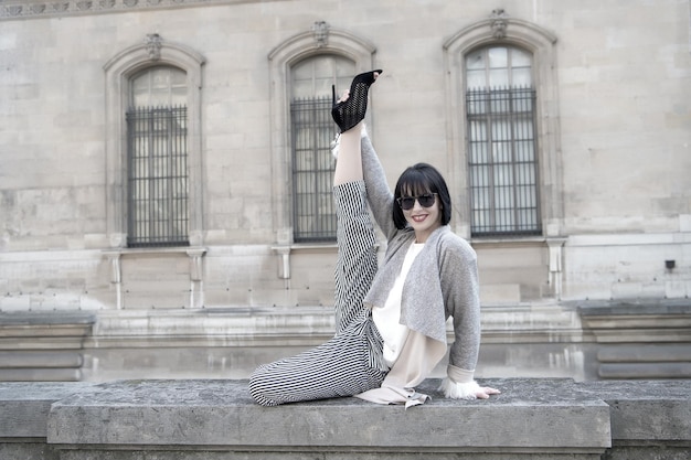 Garota feliz faz exercício de alongamento de perna dividida na cerca de pedra na fachada da casa. yoga, pilates, fitness, conceito de esporte. sorriso de mulher em óculos de sol. moda, beleza, mulher parisiense