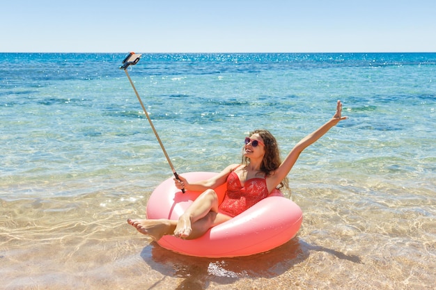 Garota feliz faz selfie em um donut polvilhado flutuando no mar sorrindo com óculos de sol para o verão