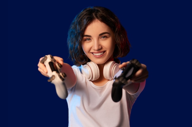 Garota feliz jogador encaracolado com dois consoles para videogame em fundo azul. jogue comigo