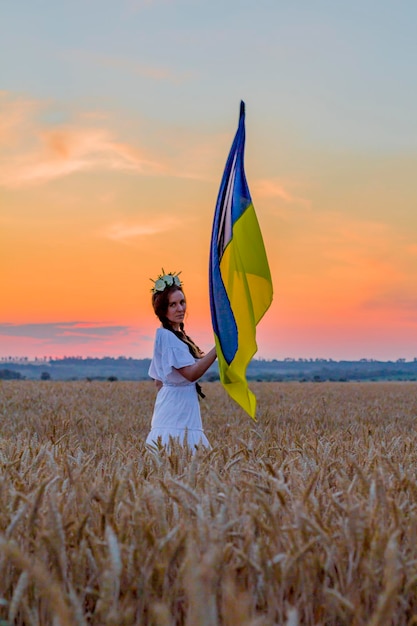 Garota feliz na camisa nacional ucraniana bordada com coroa na cabeça segura grande bandeira amarela azul contra o fundo de espigas de campo de trigo glória do dia da independência à ucrânia patriotismo