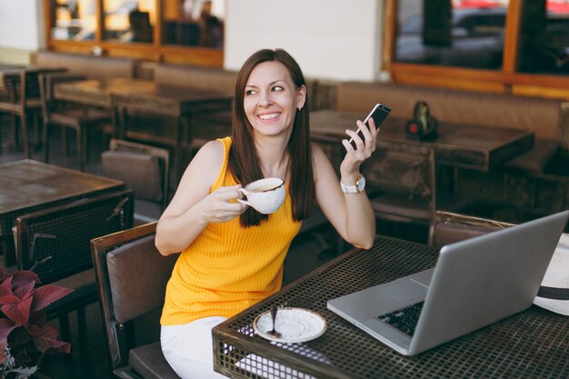 Garota feliz na rua ao ar livre, café, café, sentado à mesa com o computador laptop pc, mensagem de texto no celular, beber uma xícara de chá no restaurante durante o tempo livre