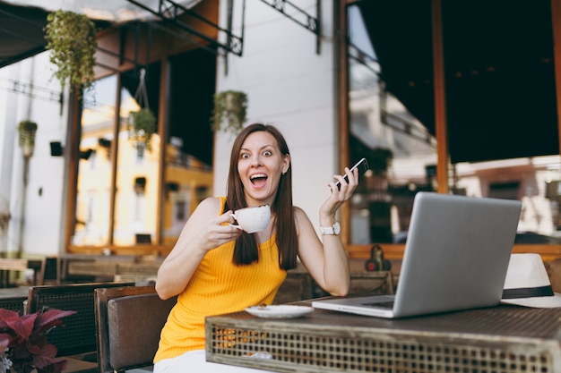 Garota feliz na rua ao ar livre, café, café, sentado à mesa com o computador laptop pc, mensagem de texto no celular, beber uma xícara de chá no restaurante durante o tempo livre