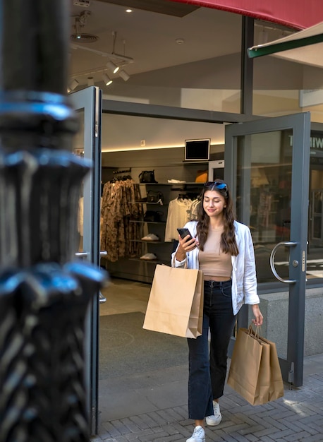 Garota feliz saindo de uma loja, celular na mão e sacolas de compras Dia de compras pechinchas sexta-feira negra