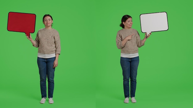 Garota feliz sorridente segurando balão de papelão para criar anúncio no estúdio, mostrando o ícone de papelão na câmera. Alegre modelo feminino com placa em pé sobre fundo greenscreen.