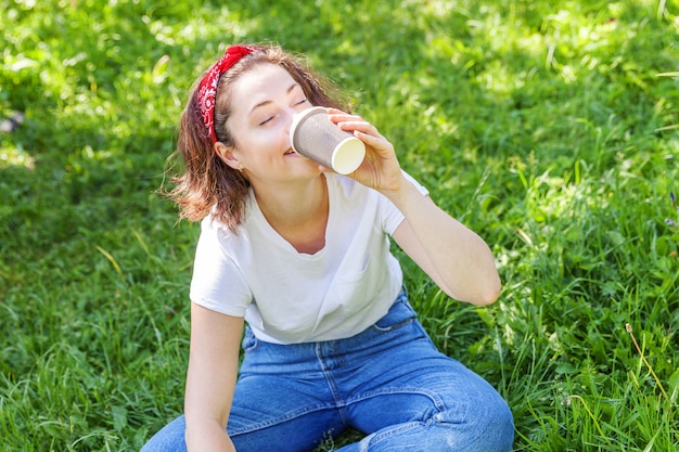 Garota feliz sorrindo ao ar livre, tendo a pausa para o almoço. A mulher nova bonita de brunete com leva embora o copo de café que descansa no parque ou na grama verde do jardim. Freelancer de educação descansando o conceito de lazer.