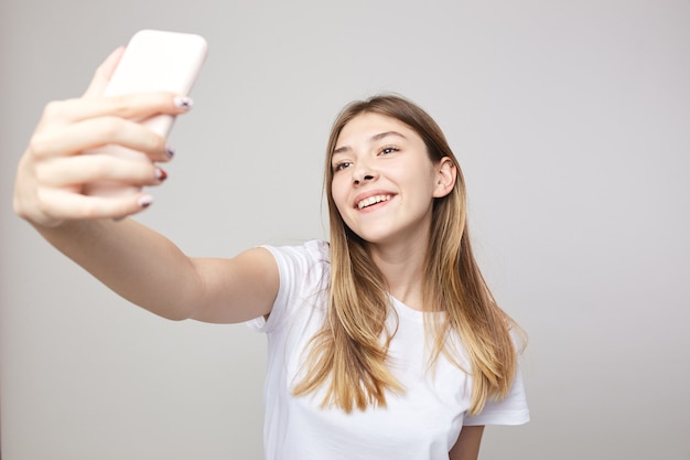 Garota feliz vestida com uma camiseta branca fazendo selfie em um fundo branco no estúdio