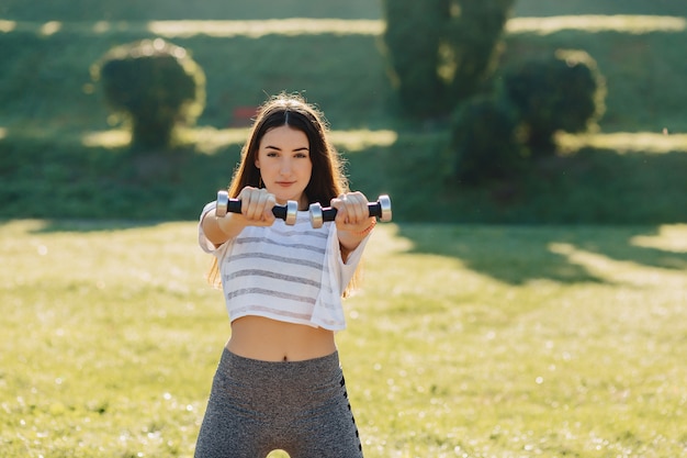 Garota fitness fazendo exercícios com halteres no pôr do sol na grama do lado de fora