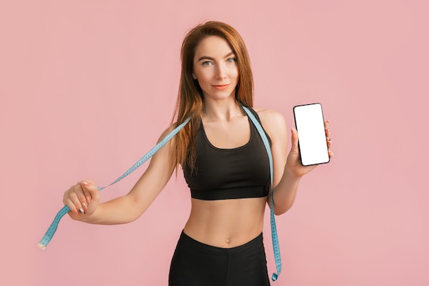 Garota fitness sorrindo e segurando um telefone com uma maquete e posando segurando a fita métrica em um sportswear preto em uma parede rosa