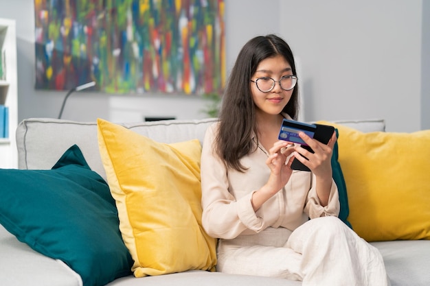 Garota focada de beleza asiática está na sala de estar no sofá nas mãos ela segura telefone e cartão