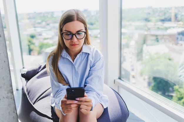 Garota freelancer feliz em óculos clássicos olhando para o telefone e sorrindo enquanto está sentado no espaço de coworking moderno mulher milenar despreocupada em óculos aproveitando o tempo de lazer para comunicação