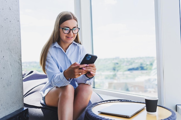 Garota freelancer feliz em óculos clássicos olhando para o telefone e sorrindo enquanto está sentado no espaço de coworking moderno mulher milenar despreocupada em óculos aproveitando o tempo de lazer para comunicação