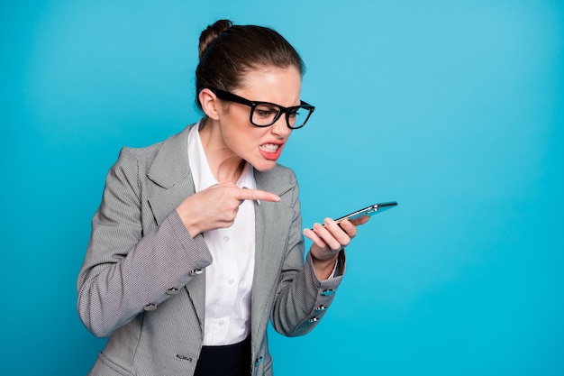 Garota furiosa executiva usando smartphone apontar dedo alto-falante usar jaqueta isolada de fundo de cor azul