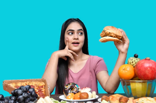 garota gastronômica sentada à mesa de frutas sorrindo, segurando um hambúrguer e olhando para ele, modelo indiano do paquistanês