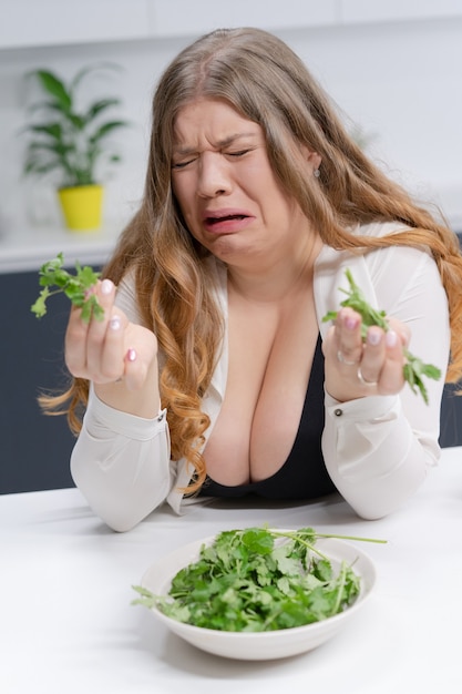 Garota gorda não feliz segurando fundição de salada fresca. Jovem de corpo curvilíneo com longos cabelos loiros sentada em uma cozinha moderna