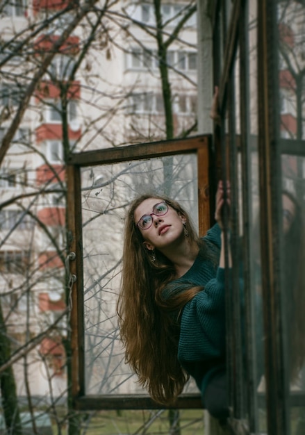 Garota graciosa com olhos inteligentes e cabelos compridos olha pela janela de madeira e caiu o frio do outono.