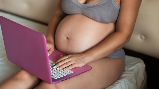 Garota grávida atraente navega no laptop e toca suavemente seu estômago. Feche as mãos da mulher no laptop com gravidez avançada de barriga grande.