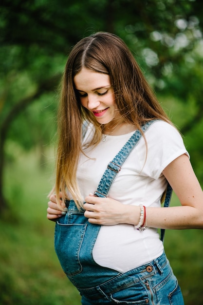 Garota grávida feliz fique de mãos dadas na barriga, fique ao ar livre na superfície do jardim com árvores verdes