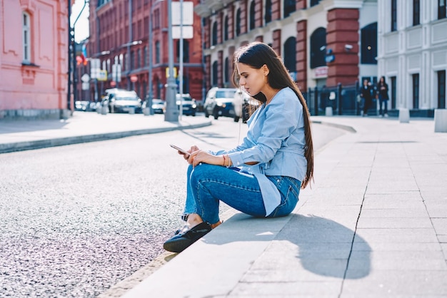 Garota hipster adolescente concentrada sentada na rua e usando o celular para blogar