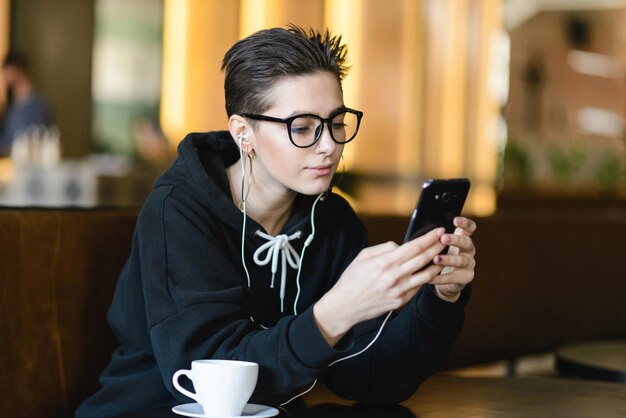 Garota hipster de atalho descansando na cafeteria ouvindo música favorita em fones de ouvido e conversando em redes