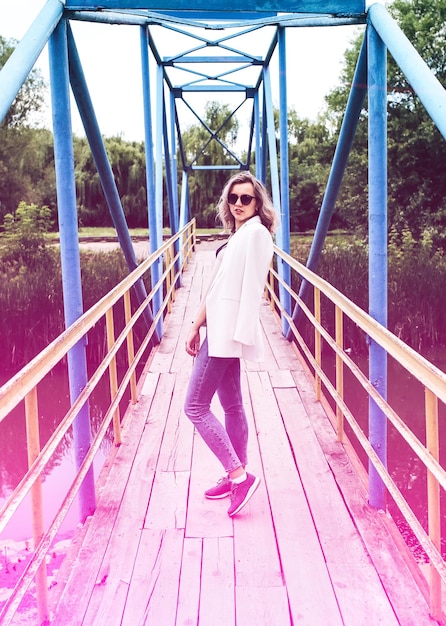 Foto garota hipster em óculos legais posando para foto de modelo jovem elegante está de pé na ponte sessão de fotos de verão na natureza ao ar livre conceito de estilo de vida