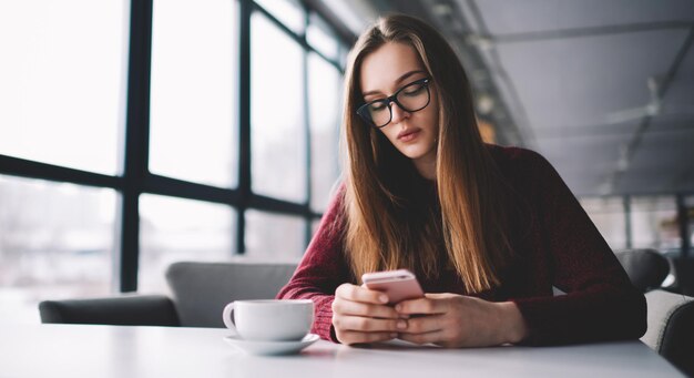 Garota hipster em óculos sentado no café universitário enviando mensagens de texto na rede social