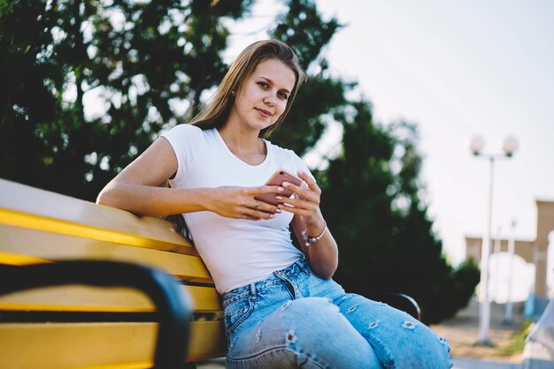 garota hipster positiva segurando o celular na mão olhe para a câmera descansando lazer no parque despreocupado