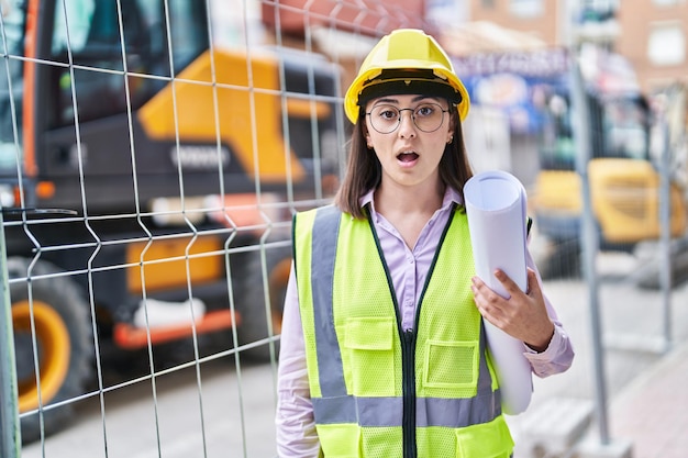 Garota hispânica usando capacete de arquiteto no canteiro de obras assustado e espantado com a boca aberta para surpresa, cara de descrença