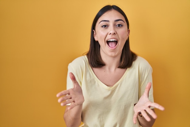 Garota hispânica vestindo camiseta casual sobre fundo amarelo comemorando louco e maravilhado pelo sucesso com os braços levantados e olhos abertos gritando animado. conceito de vencedor