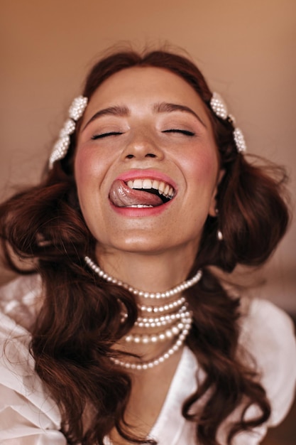 Foto garota impertinente com joias de pérolas mostra a língua e ri no fundo bege