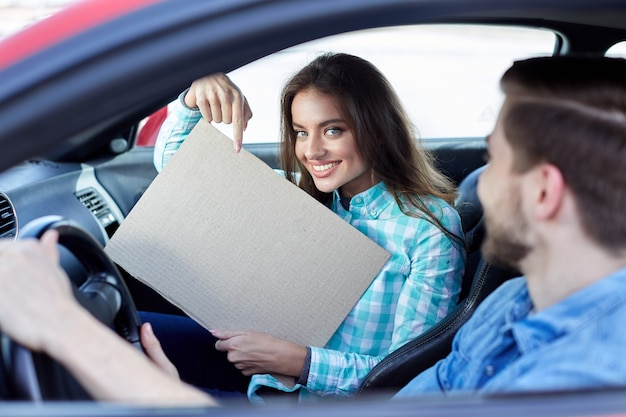 Garota incrível segurando um tablet, jovem casal sentado no carro e olhando para a câmera, sorrindo, feliz, copie o espaço, mock up, apontando no tablet.