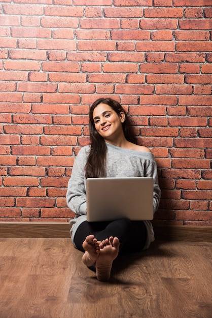 Garota indiana ou mulher sentada com um laptop ou usando um laptop sobre um piso de madeira contra uma parede de tijolo vermelho