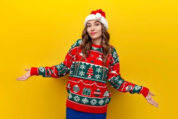 garota insegura com suéter de natal e chapéu de papai noel encolhendo os ombros em fundo amarelo