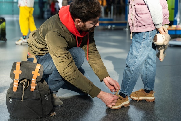 Garota irreconhecível em pé enquanto o pai amarra os cadarços dos sapatos no aeroporto antes do voo. Conceito de apoio a viagens e pais