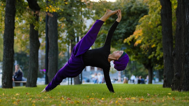 Garota islâmica esportiva forte em hijab yoga professora mulher fica na prancha lateral na grama no parque fazendo Ardhachandrasana Vasishthasana asana pose de exercício ao ar livre treino em pé bom alongamento