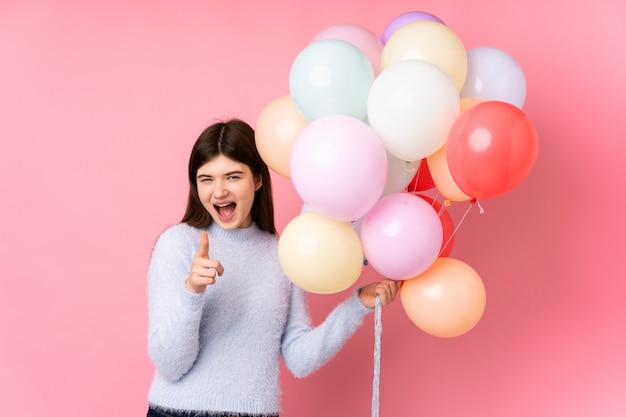 Garota jovem adolescente segurando muitos balões sobre o dedo de pontos de parede rosa para você