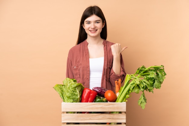 Garota jovem agricultor com legumes recém colhidos em uma caixa apontando para o lado para apresentar um produto