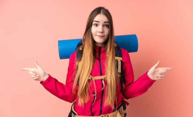 Garota jovem alpinista com uma mochila grande isolada na parede rosa, apontando para as laterais, tendo dúvidas