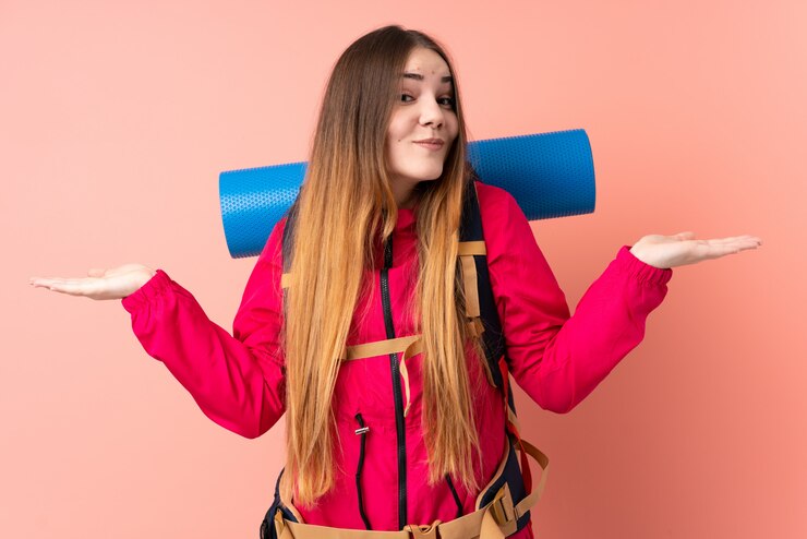 Foto garota jovem alpinista com uma mochila grande isolada na parede rosa, tendo dúvidas com a expressão do rosto confundir