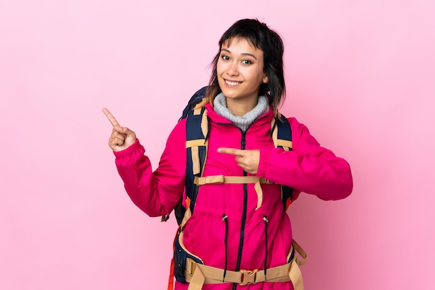 Garota jovem alpinista com uma mochila grande sobre dedo apontando rosa isolado para o lado