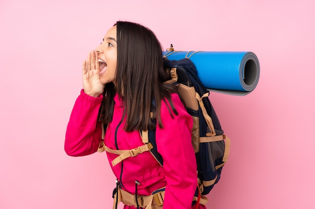 Garota jovem alpinista com uma mochila grande sobre parede rosa isolada, gritando com a boca aberta para a lateral