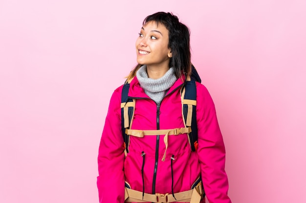 Garota jovem alpinista com uma mochila grande sobre parede rosa isolada, rindo e olhando para cima
