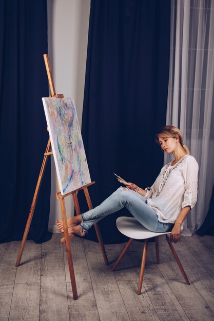 Garota jovem artista em uma camisa branca e calça jeans azul, pinta uma imagem sobre tela na oficina. um jovem estudante usa pincéis, telas e cavaletes. trabalho criativo para crianças e adultos.