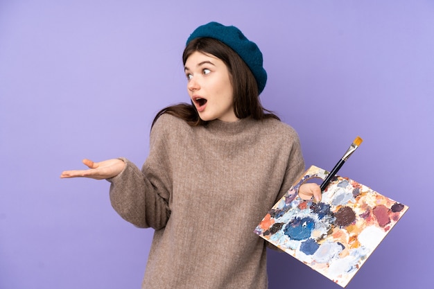 Garota jovem artista segurando uma paleta sobre parede roxa com expressão facial de surpresa
