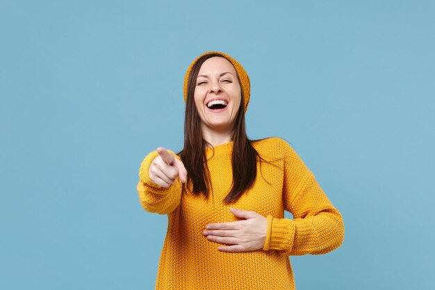 Garota jovem de suéter amarelo e chapéu posando isolado no retrato de estúdio de fundo azul a rir. Conceito de estilo de vida de emoções sinceras de pessoas. Mock up espaço de cópia. Apontando o dedo indicador na câmera.